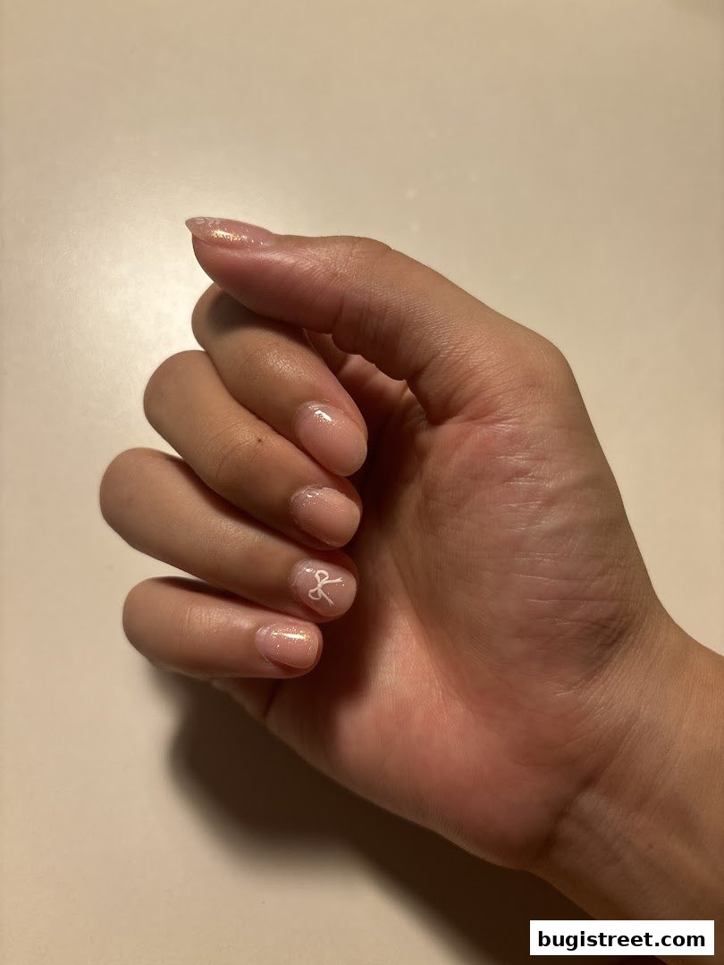 A close-up of a hand with neatly manicured nails, featuring a subtle glitter polish and a delicate white bow design.
