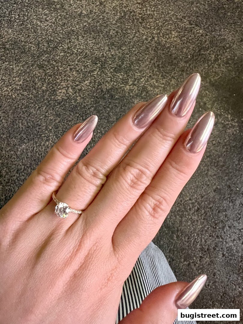 A hand with long, almond-shaped nails painted in a metallic rose gold polish, wearing a diamond ring.
