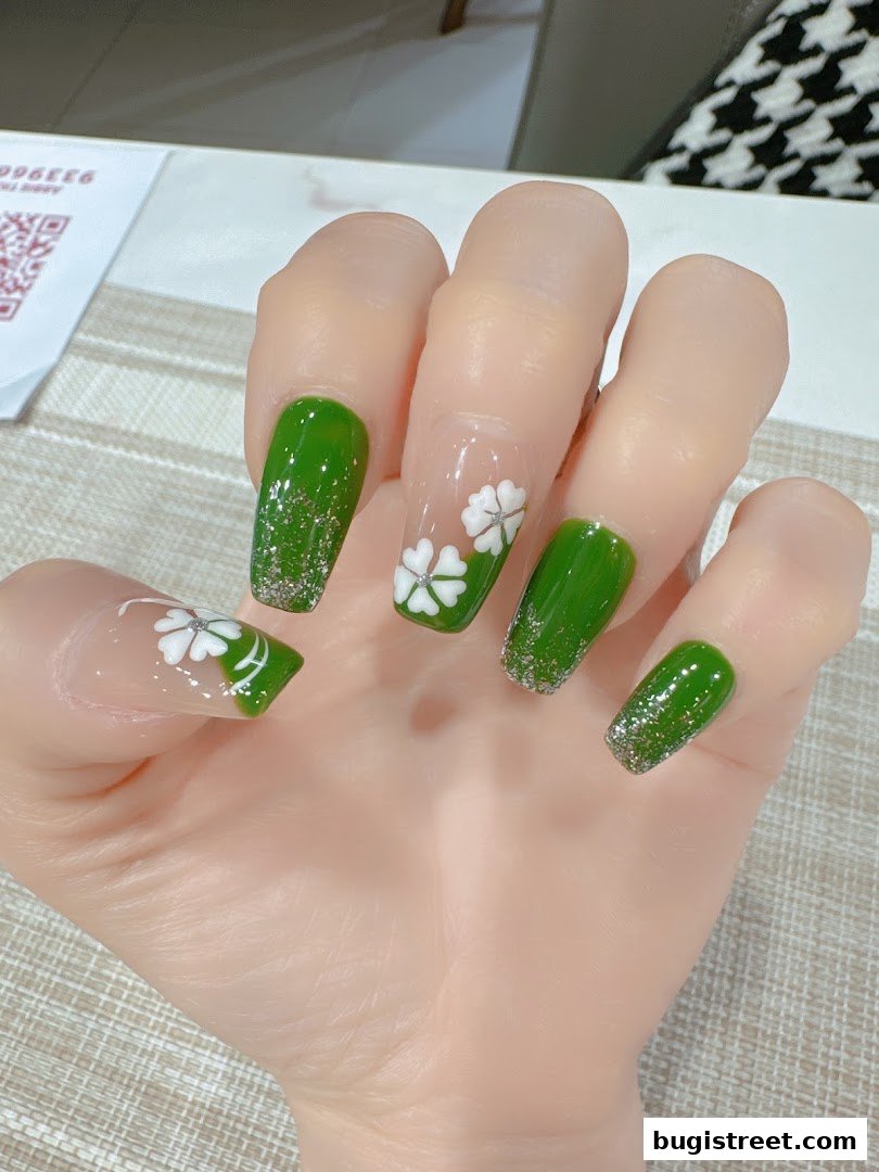 A hand with beautifully manicured green nails featuring glitter and white clover designs.