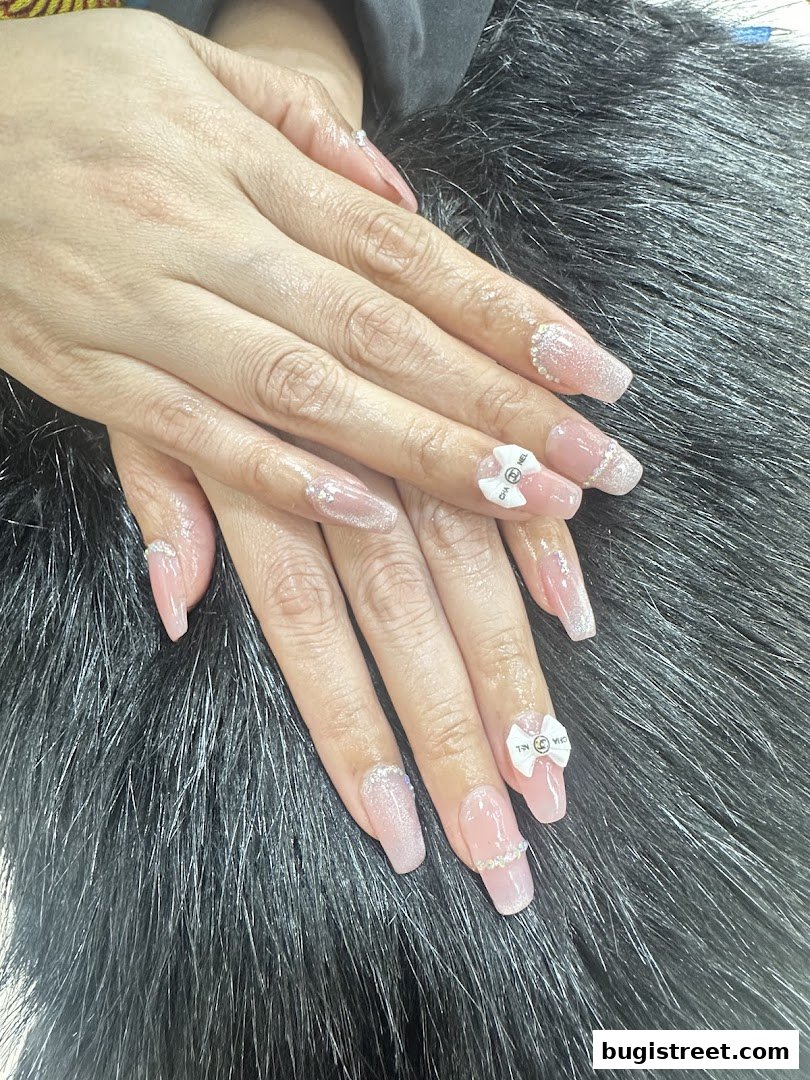 A close-up of hands with beautifully decorated, long, light pink nails featuring glitter and Chanel-inspired bows.