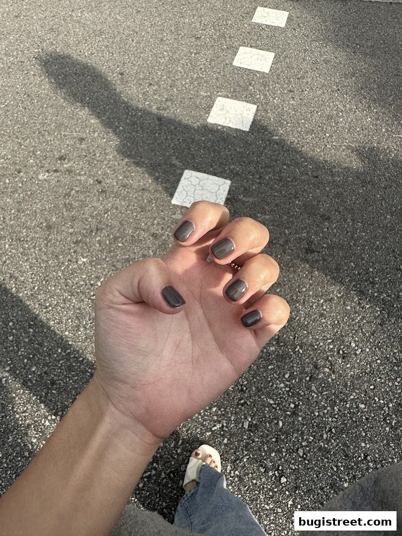 A person's hand with dark grey nail polish is shown against an asphalt background.