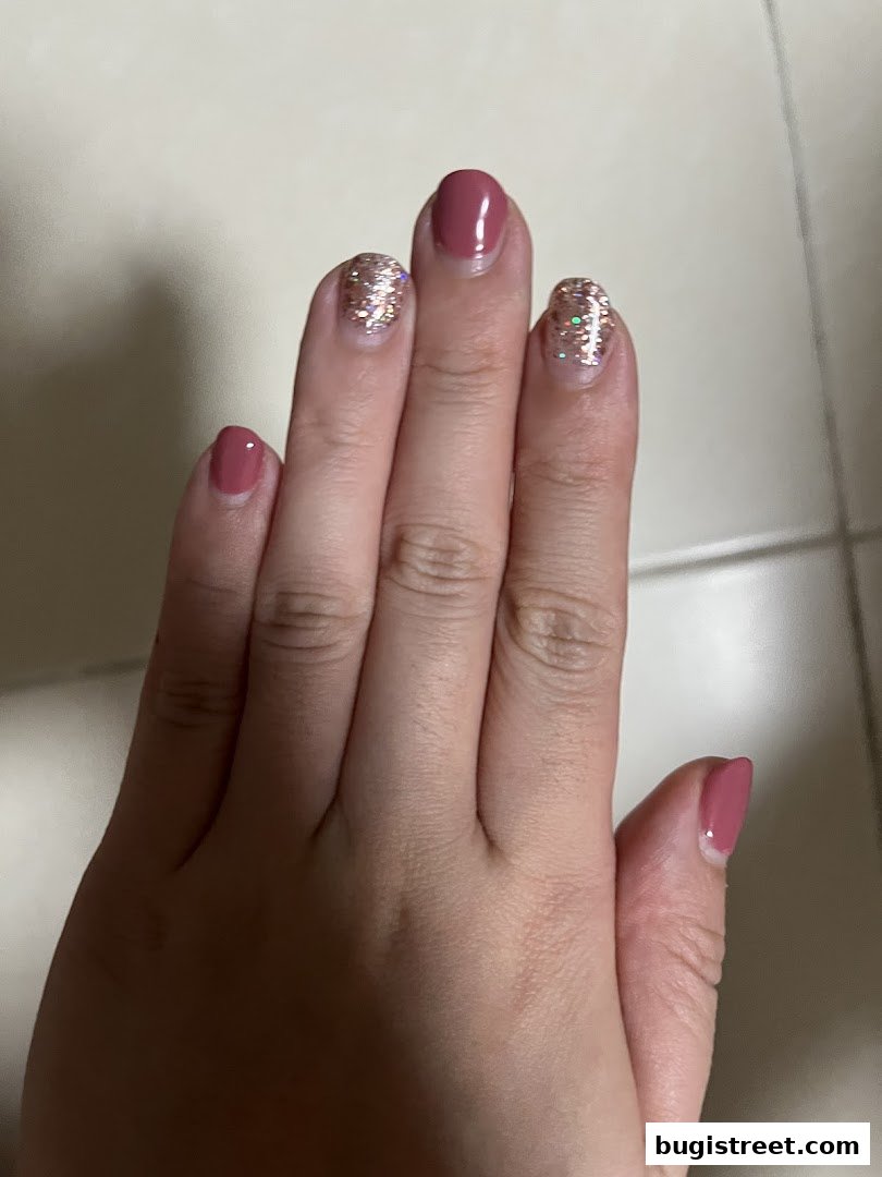A close-up of a hand with manicured nails, featuring a mix of solid rose-gold polish and glitter accents.