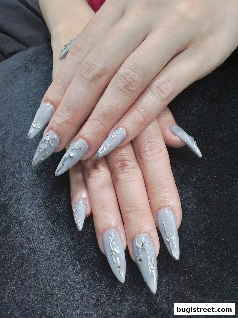Close-up of hands with elegant grey stiletto nails adorned with delicate silver designs and rhinestones.