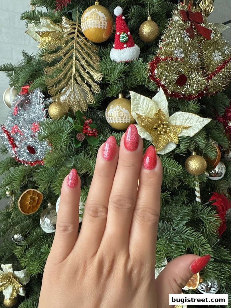 A hand with sparkling red nails is shown against a festive Christmas tree background.