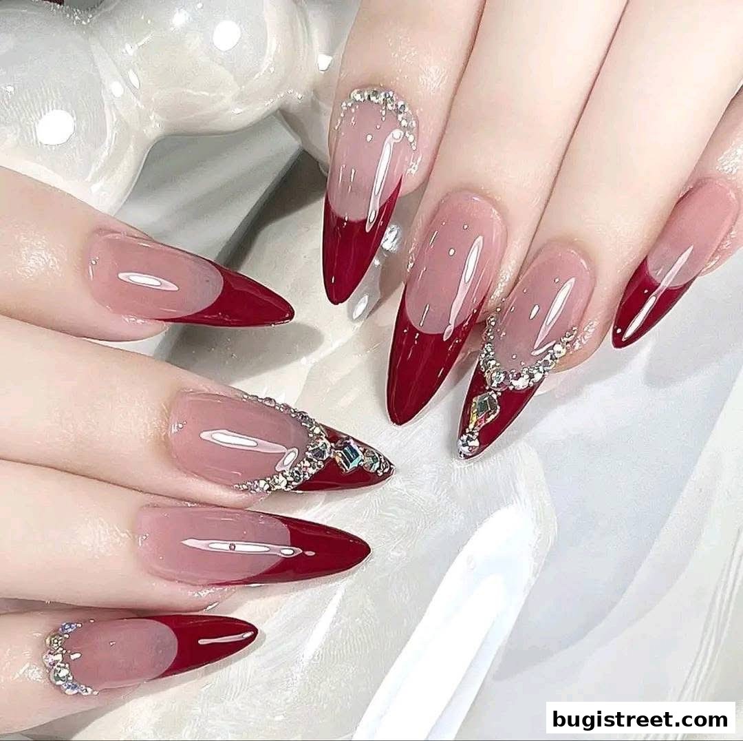 Close-up of hands with elegant, long, stiletto-shaped nails featuring a deep red French tip and intricate rhinestone embellishments.