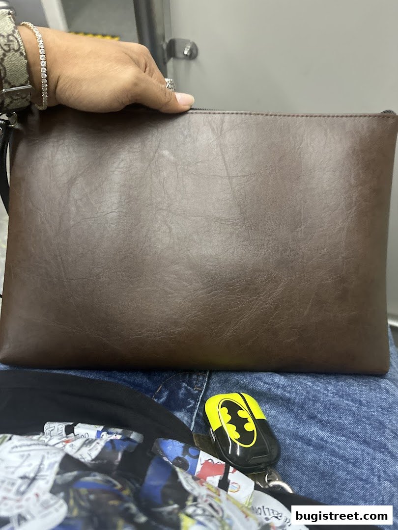 A person holds a brown clutch bag with a Batman-themed keychain and a bracelet.