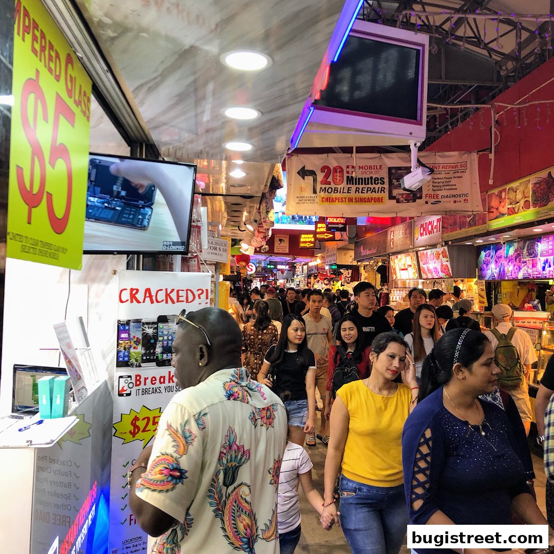 A busy market street with shops selling phone repairs and accessories, with many people walking by.
