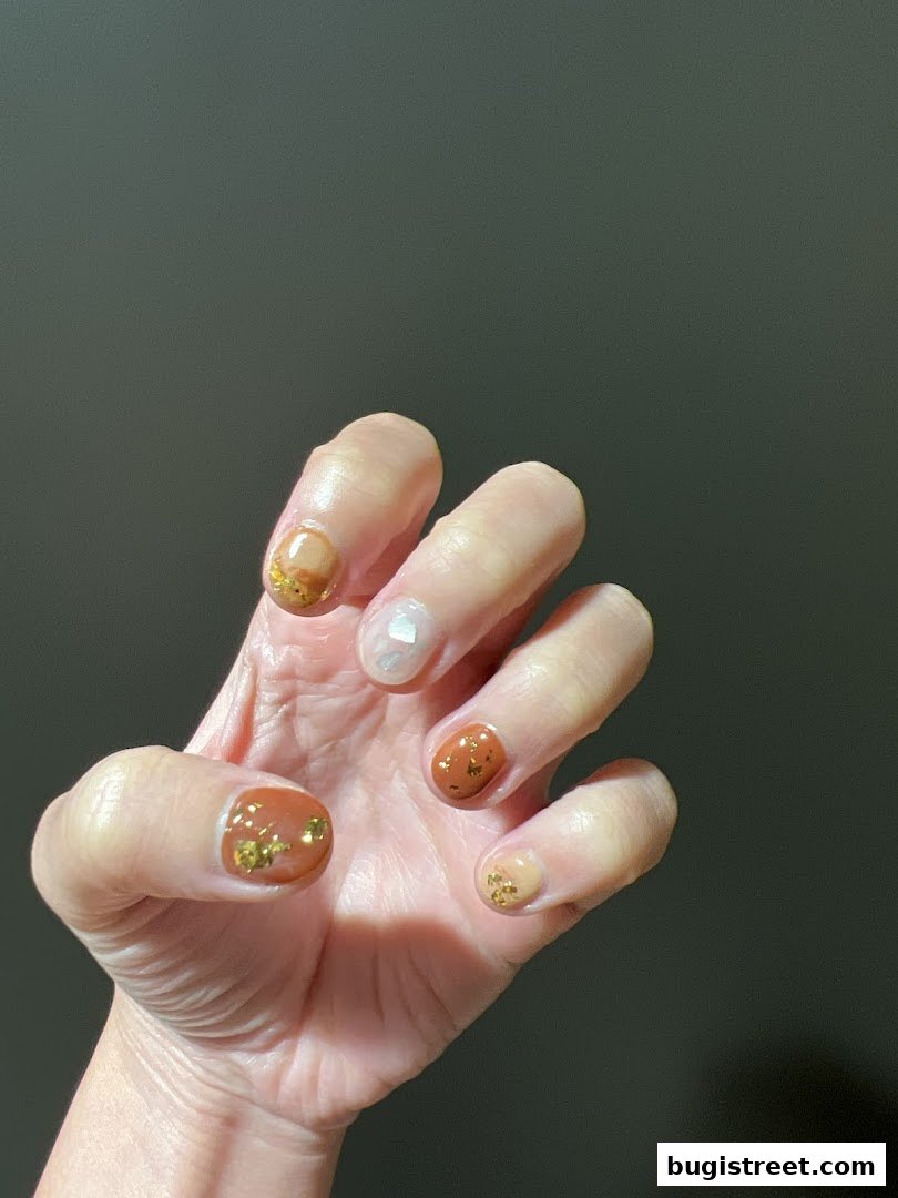 A hand with beautifully designed nails featuring a mix of brown, beige, and pearl colors with gold flakes.