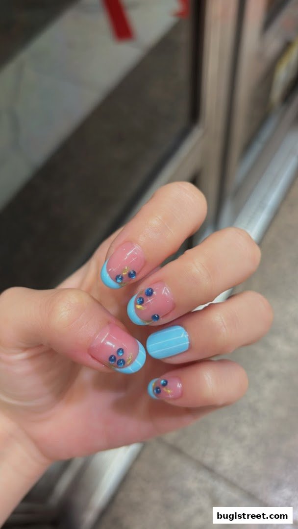 Close-up of a hand with beautifully decorated French tip nails featuring blue gems and gold accents.