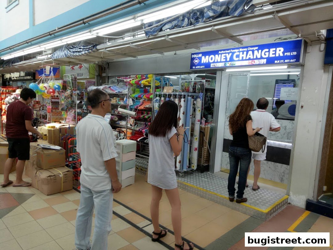 People are seen at the entrance of a Dollar Money Changer shop with various goods displayed.