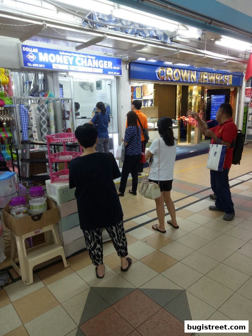 People are queuing outside a money changer and a jewelry store in a mall.