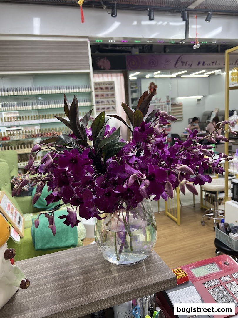 A vase of purple orchids sits on a counter in a nail salon, with shelves of nail polish visible.