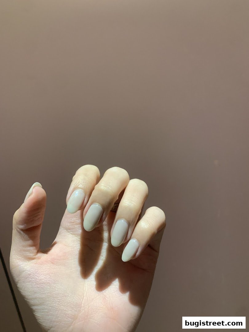 A hand with neatly manicured, light beige oval-shaped nails against a plain background.