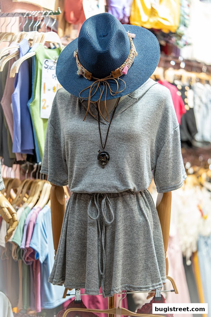 A mannequin displays a grey dress and a blue hat, with clothing racks in the background.