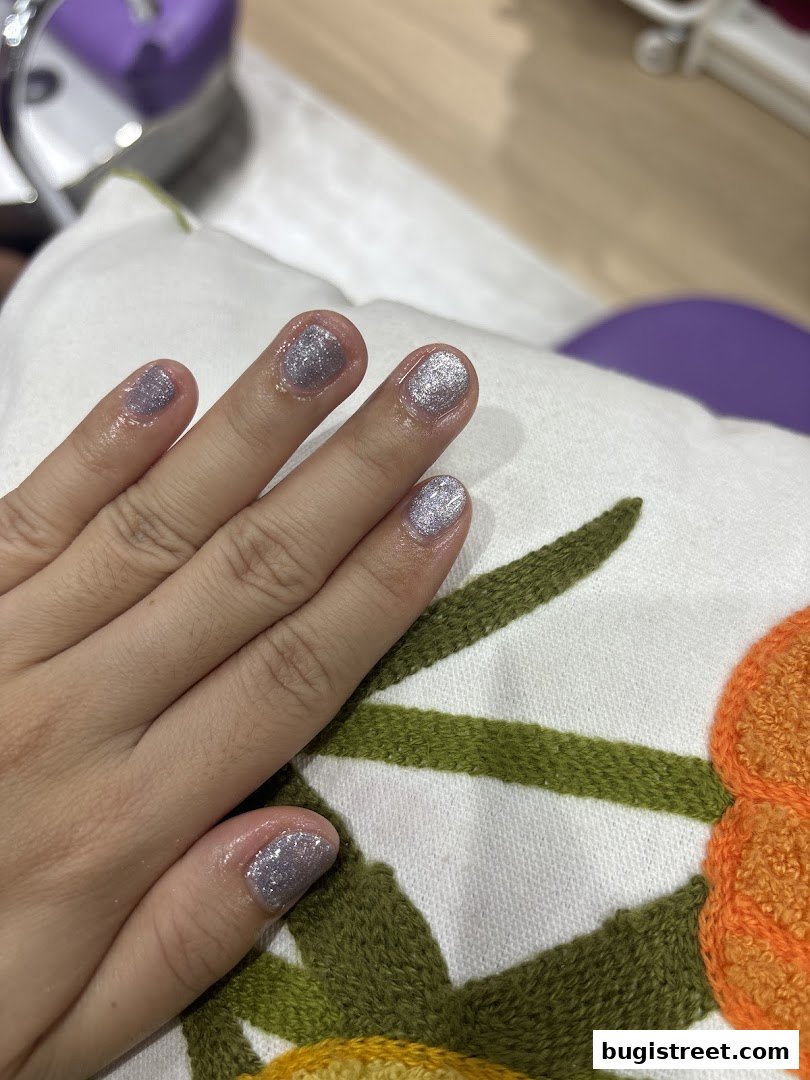 A close-up of a hand with freshly painted glittery silver nails resting on a white cushion with floral embroidery.