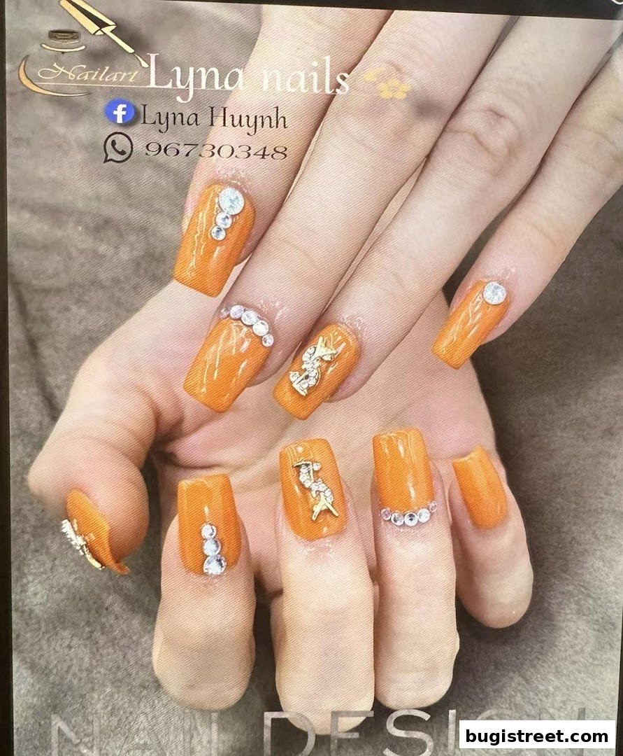 Close-up of hands with vibrant orange acrylic nails adorned with rhinestones and charms.