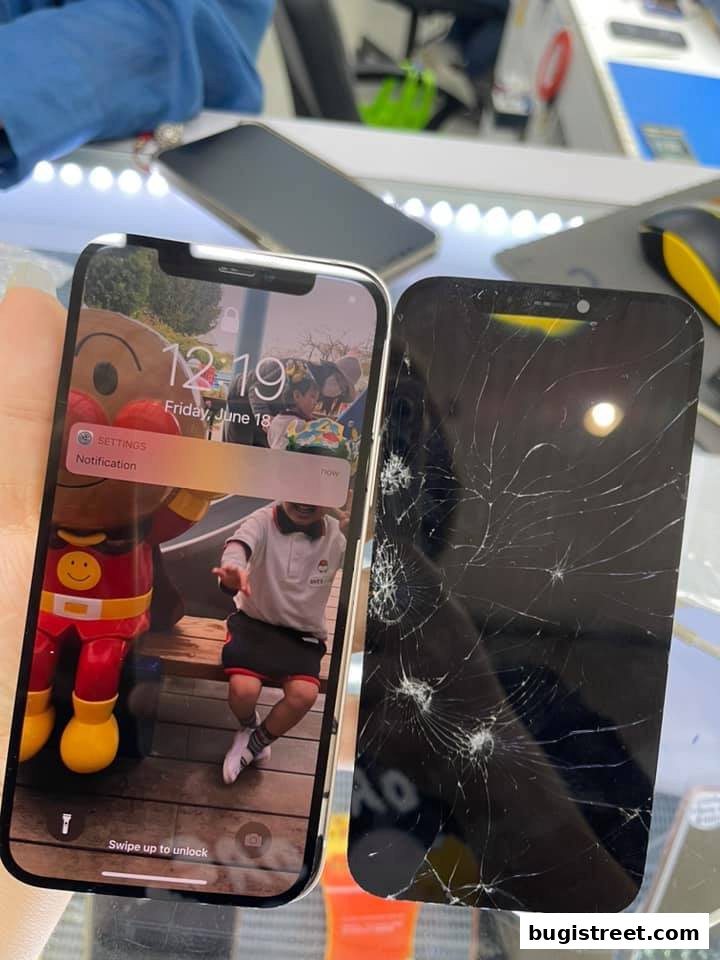 A hand holds a working iPhone next to a phone with a severely cracked screen.