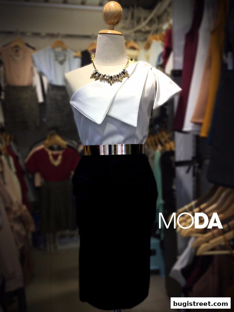 A mannequin displays a white one-shoulder top and black skirt with a gold belt.