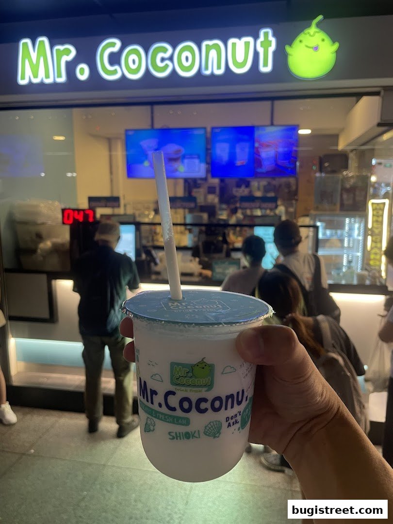 A hand holds a Mr. Coconut drink in front of the shop's illuminated sign.