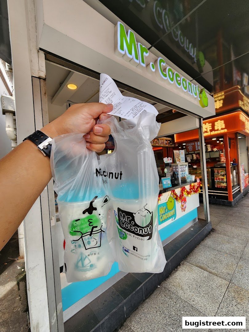 A hand holds two Mr. Coconut drinks in a plastic bag outside the shop.