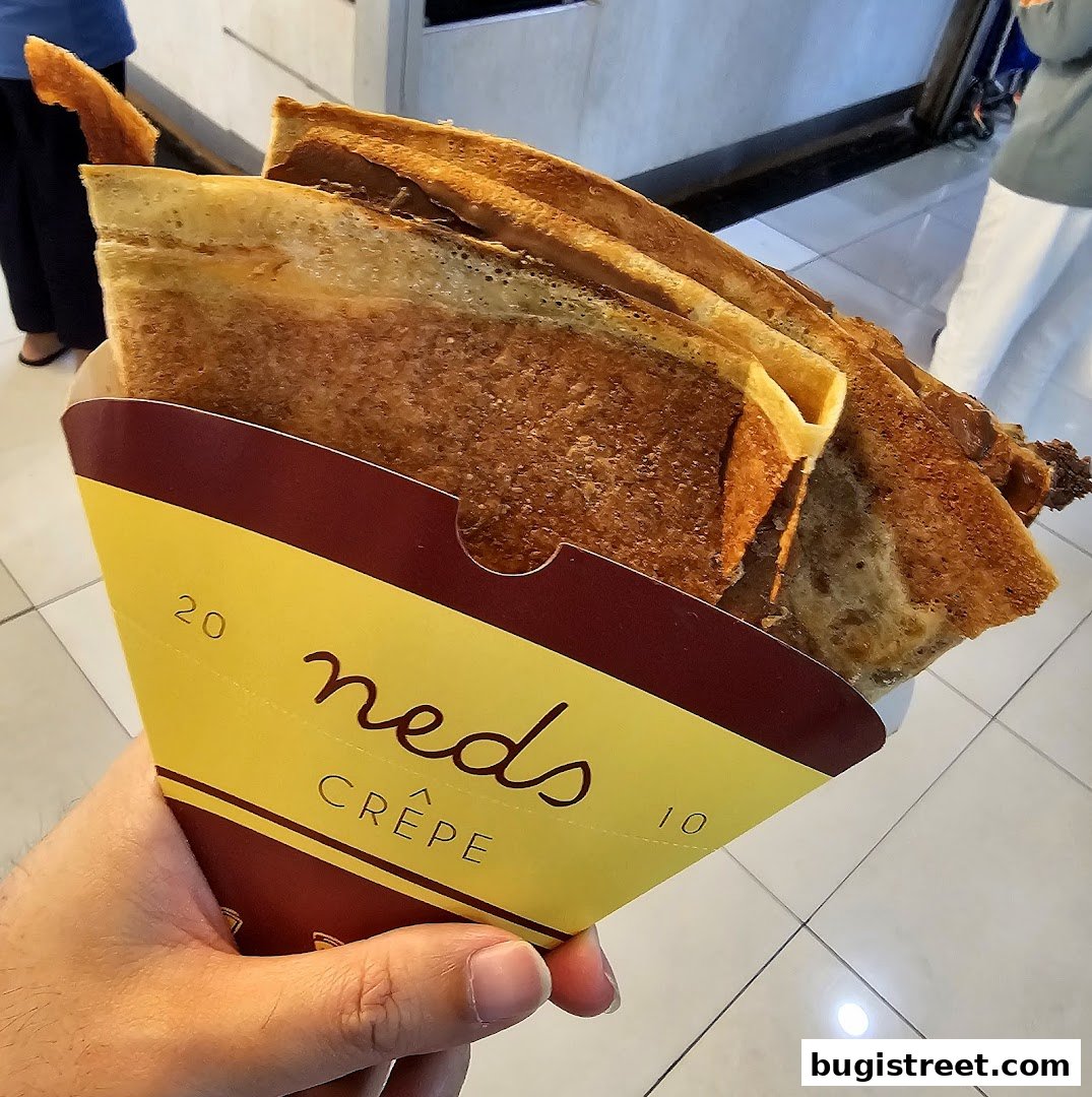 A hand holds a freshly made crepe in a branded paper holder.