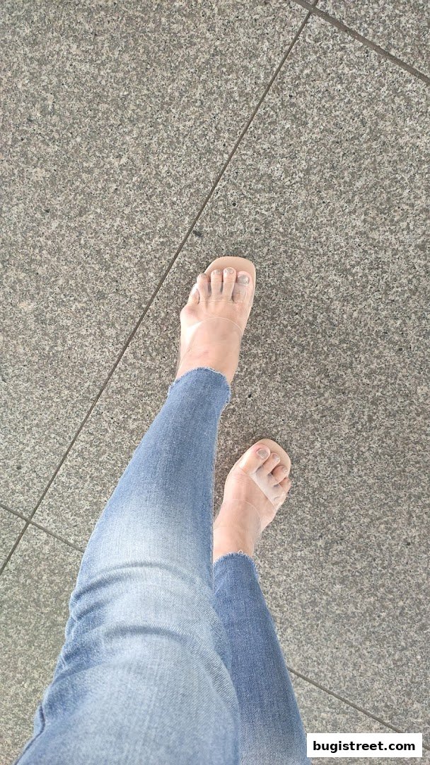 A person's feet wearing clear sandals and blue jeans on a granite floor.