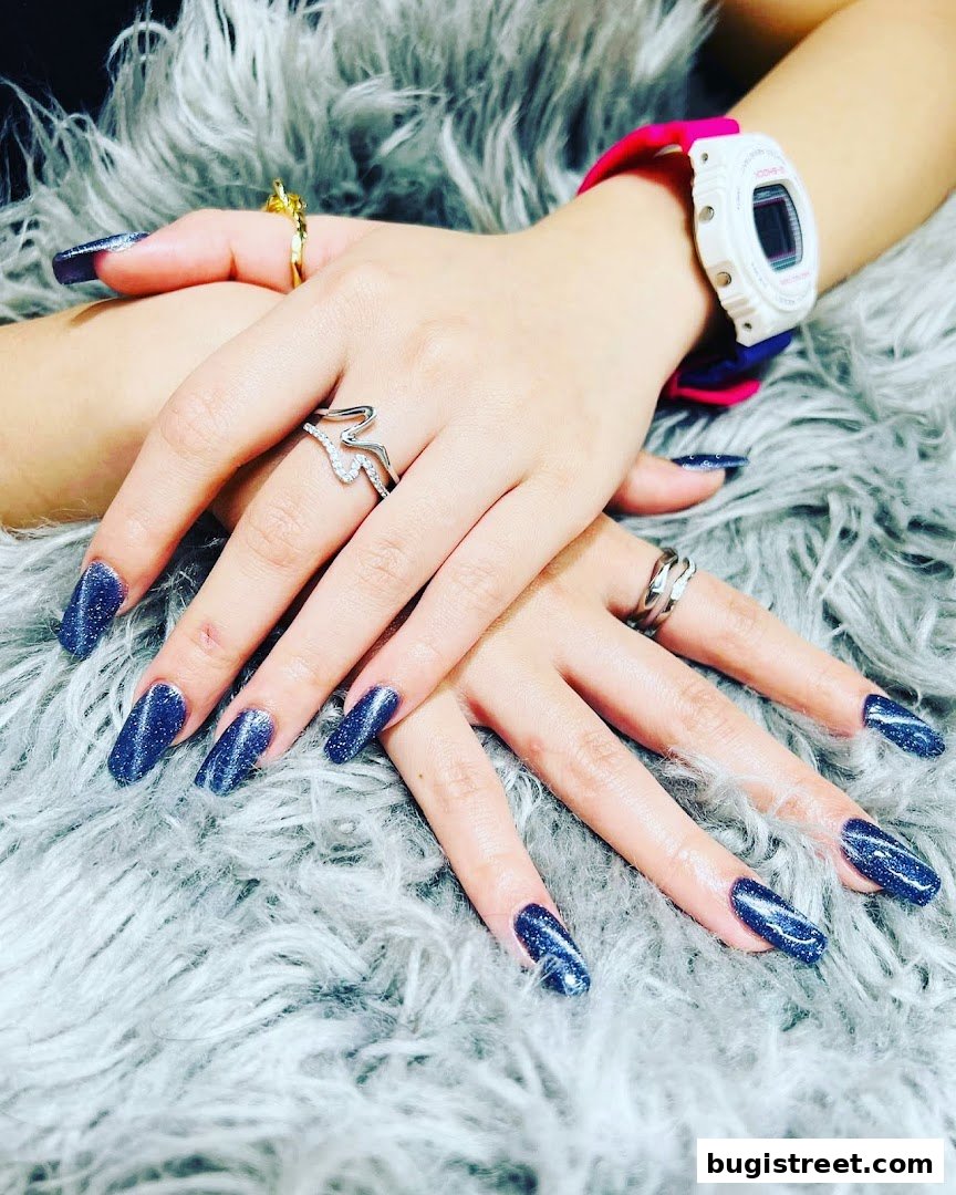 Close-up of hands with long, sparkly dark blue nails and rings.