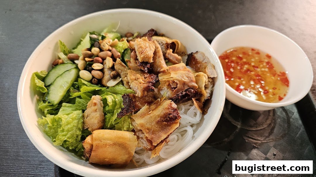 A bowl of vermicelli noodles with grilled meat, vegetables, peanuts, and a side of dipping sauce.