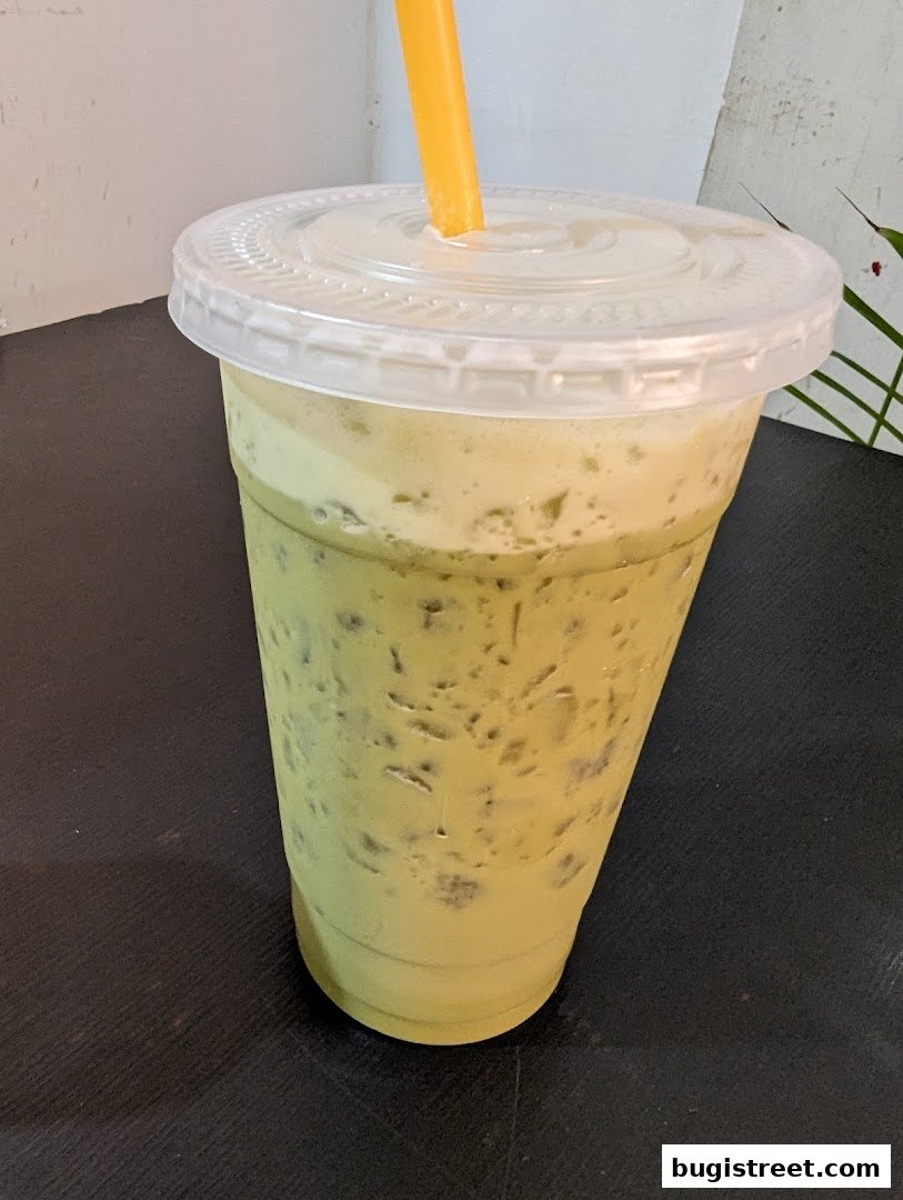 A tall, clear plastic cup filled with a creamy green beverage and ice, topped with a lid and a yellow straw.
