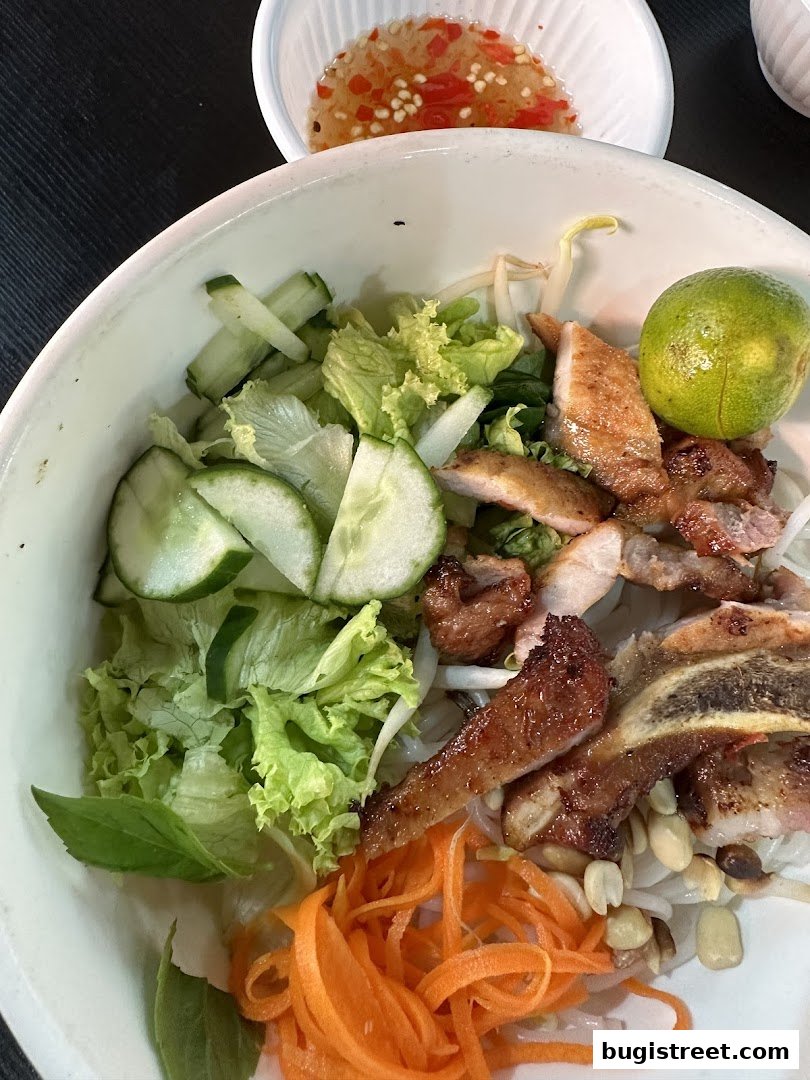 A close-up of a delicious bowl of vermicelli noodles with grilled pork, fresh vegetables, and a side of dipping sauce.