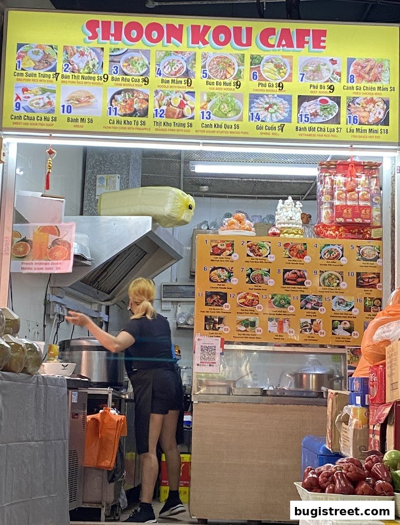 A food stall with a large menu board displaying various Vietnamese dishes and a staff member preparing food.