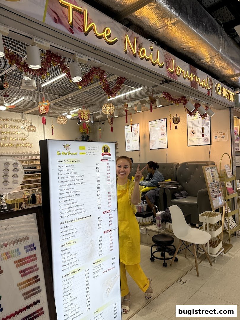 A smiling woman in a yellow apron stands in front of 'The Nail Journal' shop, displaying a menu.