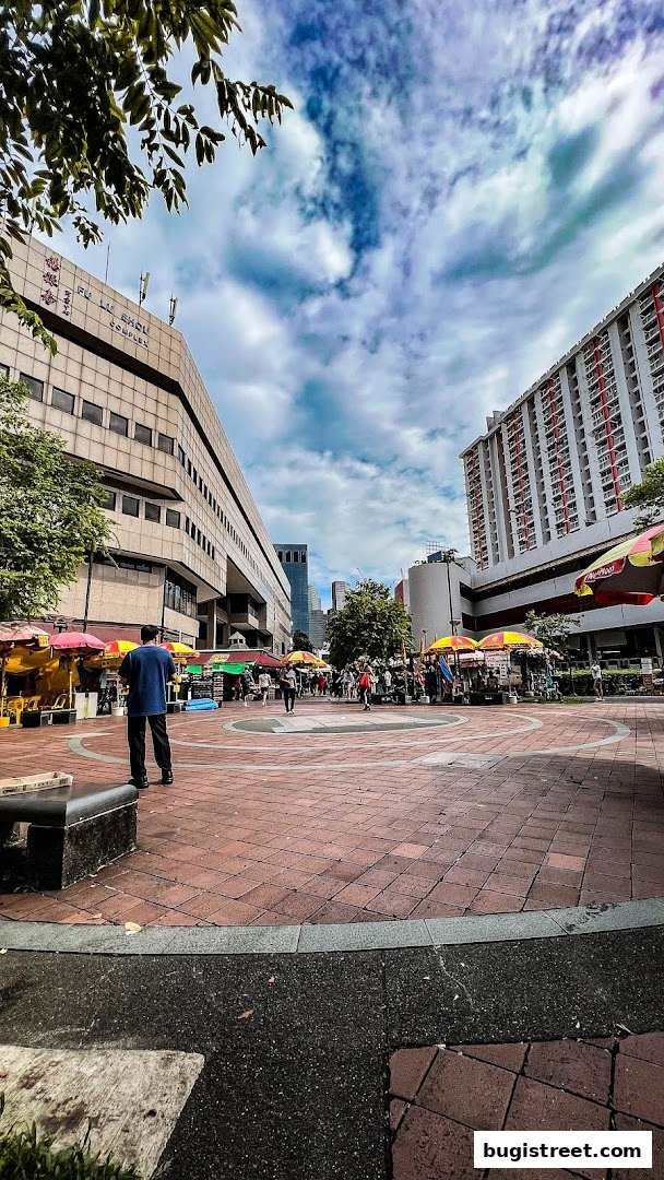An outdoor plaza with market stalls and people under a cloudy sky.